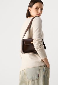 Brun skinn crossover-veske med klaffelukking og gullfarget hardware, båret over en lys beige genser med ribbestrikkede mansjetter, kombinert med lyse denim.