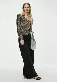 Woman wearing one-shoulder sequined top, wide black pants, black sandals, holding silver handbag, standing against plain background.