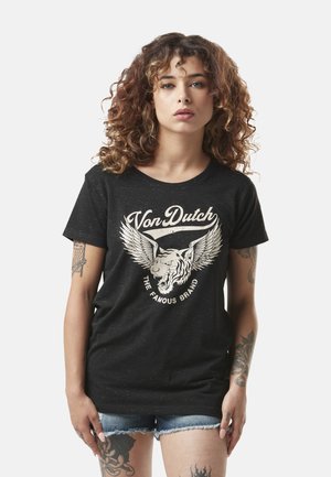 Jeune femme aux cheveux bouclés portant un t-shirt noir Von Dutch avec un graphisme de tigre ailé et un short en jean, exhibant plusieurs tatouages.