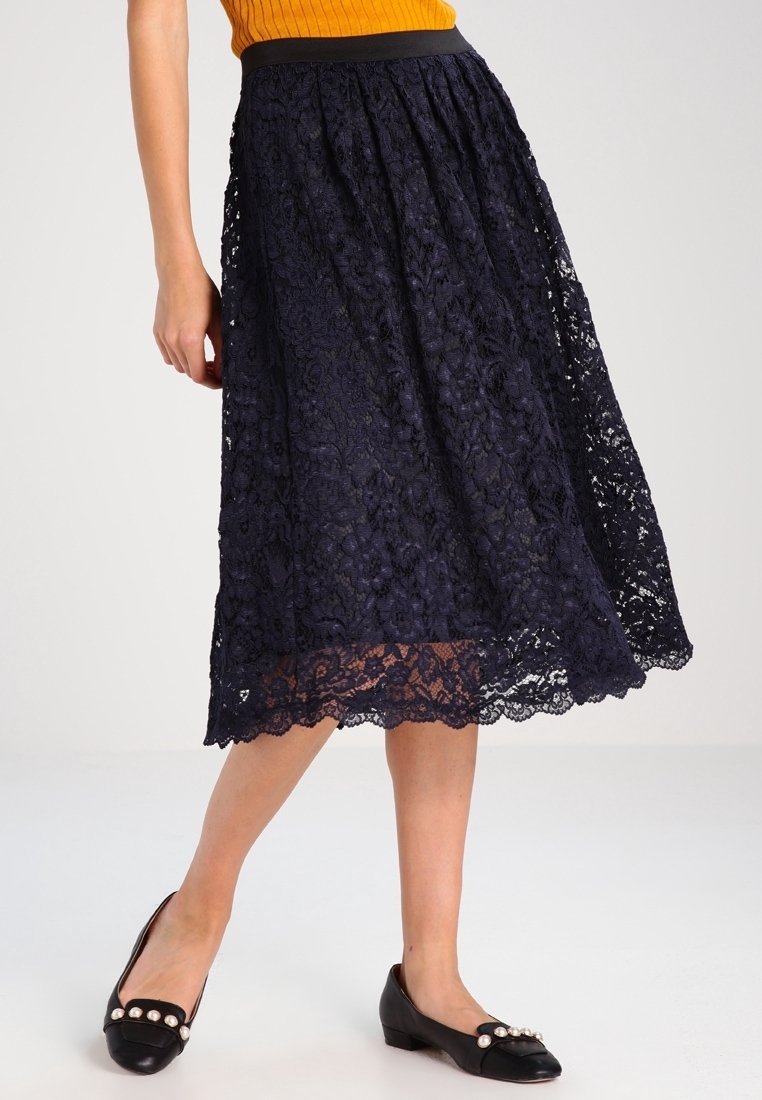 Jupe midi en dentelle navy avec une taille froncée, ornée de motifs floraux et d'un ourlet en dentelle. Associée à des ballerines noires embellies de perles.