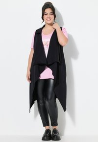 Schwarzes ärmelloses Oberteil über einem rosa T-Shirt mit glitzerndem Text, kombiniert mit glänzenden schwarzen Leggings und klobigen schwarzen Schuhen. Glatte Stoffe, lockerer Schnitt.