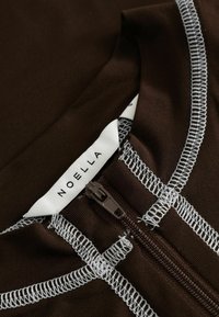 Καφέ ρούχο με εμφανή λευκή ραφή overlock, καφέ κλειστό φερμουάρ και λευκή ετικέτα ρούχου με την επιγραφή "NOELLA" μέσα στον γιακά.