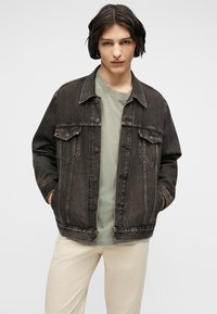 Jeune personne aux cheveux courts et foncés portant une veste en jean noire par-dessus une chemise vert clair et un pantalon beige, les mains dans les poches de la veste.