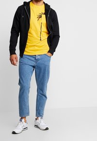 Černá mikina na zip přes žluté tričko s grafikou palmy, doplněná světle modrými džíny a bílými teniskami s námořnickými akcenty.