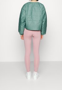 Grön kviltad bomberjacka, kortad design, som matchas med rosa leggings. Vita sneakers med guldaccenter fulländar outfiten. Släta texturer.