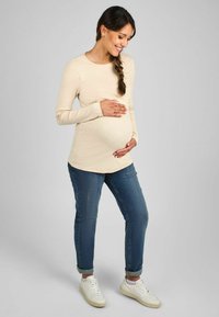 Beige garšīga piedurkņu krekls ar apaļu kaklu, apvienots ar cieši pieguļošiem ziliem džinsiem un baltiem čībām. Modelis tur grūtniecības vēderu.