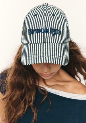 Žena s dlhými vlasmi hľadí dolu, má na sebe námornícku bielo-modrú pruhovanú čiapku s výšivkou „Brooklyn“ a námornícky top, ktorý odhaľuje jedno plece.