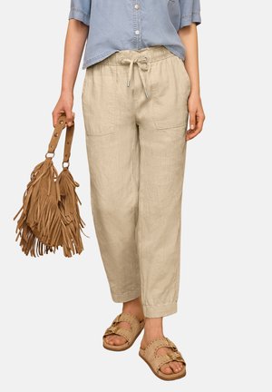 Kvinde iført beige bukser med snor, lyseblå kortærmet skjorte, brune sandaler, holder en brun taske med frynser ved siden af sig.