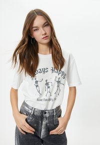 Hvid kortærmet t-shirt med grafik af dansende skeletter og tekst. Parret med højtaljede, slidte sorte jeans.