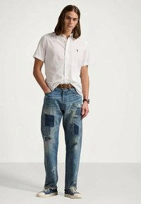 Jovem com cabelo comprido, vestindo uma camisa branca de manga curta abotoada, calças jeans azuis com remendos, cinto castanho e ténis azuis, de pé com as mãos nos bolsos.