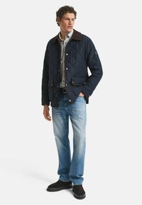 Gestepptes marineblaues Jackett mit braunem Kragen, zwei Fronttaschen und Knopfverschluss, kombiniert mit hellblauen Jeans und schwarzen Schuhen.