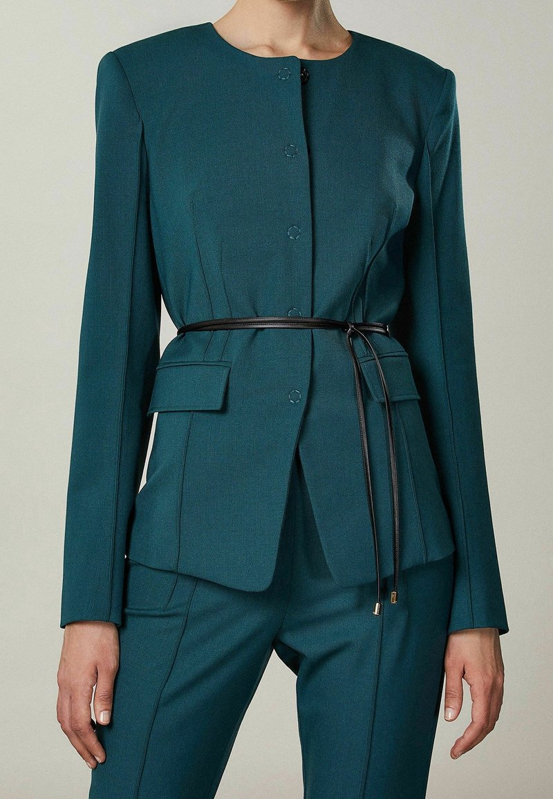 Femme portant un costume ajusté bleu sarcelle avec un col rond, une fermeture à boutons dissimulée, des poches à rabat et une fine ceinture noire nouée à la taille.