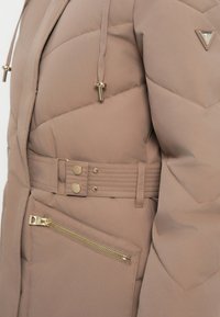 Chaqueta acolchada beige con cinturón, bolsillo con cremallera, snaps de botones dorados, cordones con puntas doradas y parche con logo triangular en la manga.