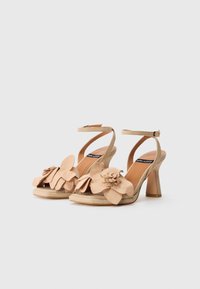 Beige hoge hakken sandaal met bloemaccenten, gemaakt van suede, open teen, enkelbandje en een blokhakdesign. Geweven bloemdetails op de bovenkant.