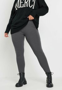 Leggings gris à côtes associés à un pull noir surdimensionné arborant le mot "MERCI" en blanc, complétés par des bottes noires à lacets.