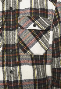 Camicia in flanella a quadri nei colori bianco, nero, verde, giallo e rosso. Presenta una tasca sul petto, bottoni neri e una texture morbida.