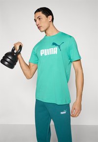Τιρκουάζ μπλουζάκι με λευκό λογότυπο "PUMA" και μαύρο kettlebell. Καθημερινό σχέδιο, κοντά μανίκια, λεία υφή. Τιρκουάζ παντελόνια με λεπτομέρεια λογότυπου.