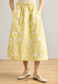 Jupe midi à motif floral jaune avec une taille élastique et des poches, fabriquée en tissu texturé. Portée avec des chaussures plates beiges.