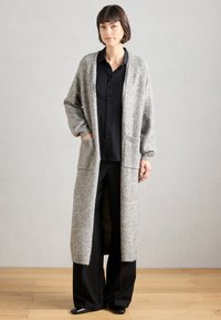 Largo cárdigan gris hecho de lana texturizada, con un frente abierto, dos bolsillos delanteros y puños acanalados, combinado con una camisa negra de botones y pantalones.