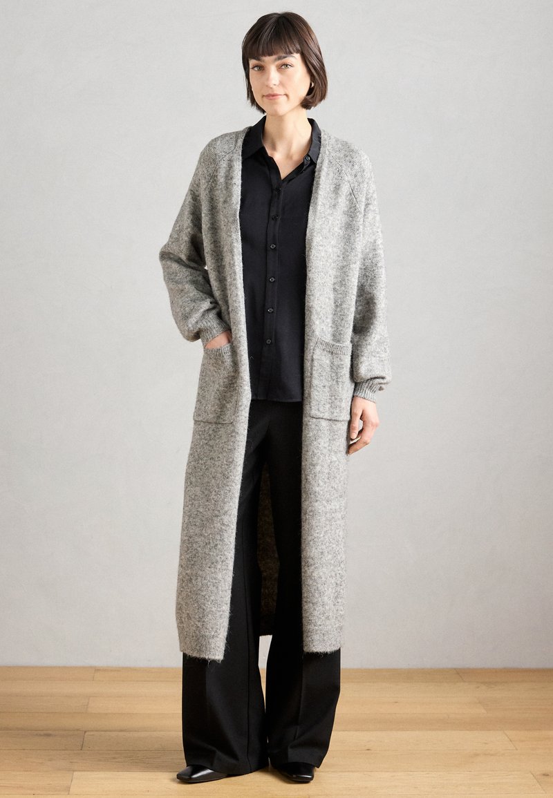 Largo cárdigan gris hecho de lana texturizada, con un frente abierto, dos bolsillos delanteros y puños acanalados, combinado con una camisa negra de botones y pantalones.