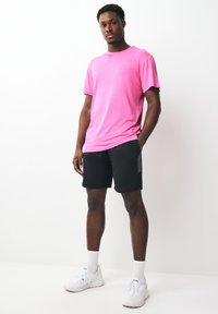 Uomo in piedi con le mani nelle tasche, indossa una maglietta rosa brillante, pantaloncini neri, calzini bianchi alti e scarpe da ginnastica bianche.