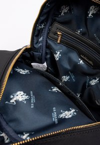 Svart interiör i en väska med gyllenblixtlås, som har ett marinblått foder med upprepade vita U.S. Polo Assn. logotyper och en ficka med blixtlås.
