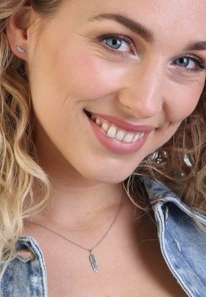 Zilveren veer hangerketting op een fijne ketting, gedragen over een denimjasje. Krullend haar en minimale make-up zichtbaar.