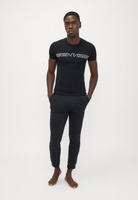 Melnā pieguļošā T-kreklā ar baltu "Emporio Armani" logo un melnās sašaurinātās sporta biksēs, abas izgatavotas no mīkstas, vieglas auduma.