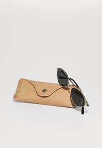 Gafas de sol de forma cat-eye en color negro con montura de metal dorado, colocadas sobre un estuche de piel texturizada de color marrón claro que presenta un cierre a presión y detalles de logotipo.