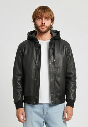 Uomo con capelli castano chiaro che indossa una giacca di pelle nera con cappuccio, maglietta bianca e jeans azzurri, in piedi davanti a uno sfondo neutro.