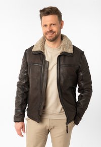 Bruine leren jas met een gestructureerde afwerking, voorzien van een beige nepbontkraag en contrasterende zwarte ritsen, gecombineerd met een beige ronde hals trui.