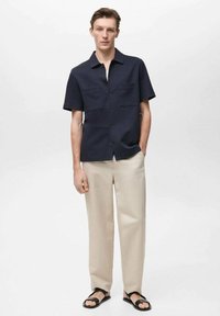 Marineblauwe korte mouwen shirt met twee voorkruimels, beige broek en zwarte sandalen. Gladde stof met een knoopsluiting en een relaxte pasvorm.