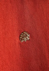 Červená látka s pletenou strukturou má na levé straně malý hnědý vyšitý design mušle, včetně detailních linií a vzorů.