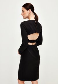 Abito nero con maniche lunghe, collo alto, scollatura sulla schiena e dettaglio vita arricciata. Texture liscia e silhouette aderente.