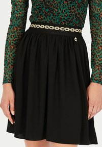 Czarna spódnica z rozkloszowanym dołem, z elastycznym pasem, ozdobiona złotym paskiem w kształcie łańcucha i małą koroną. Gładka tekstura, długość do kolan.