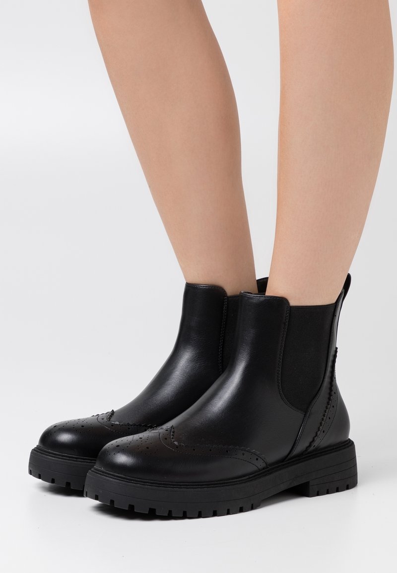 Bottes basses en cuir noir avec détails brogue et semelles épaisses portées sur des jambes nues contre un fond blanc uni.