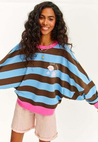 Pruhovaná oversized mikina v modré a hnědé barvě s růžovými detaily, s grafikou mušlí a textem "Hledač mušlí." V kombinaci s růžovými šortkami.