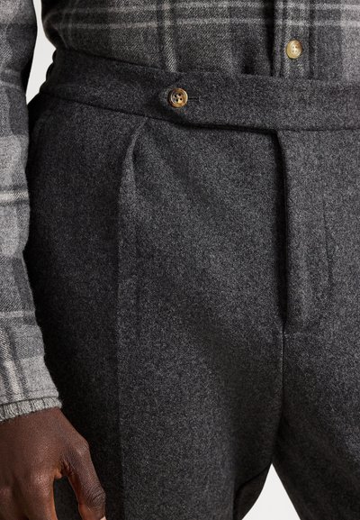 Ciemnoszare wełniane spodnie z wysokim stanem, plisami i brązowym zapięciem na guzik. Tkanina ma miękką fakturę.