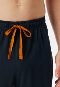 Tumman siniset loungemut ovat varustettu joustavalla vyötäröosalla, jossa on litteä oranssi nauha. Materiaalin pinta näyttää pehmeältä ja sileältä.
