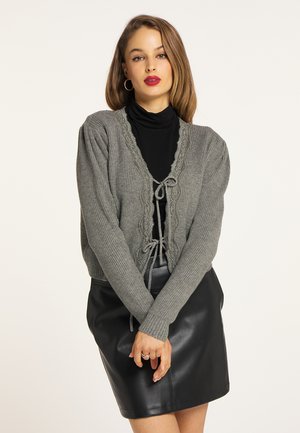 Grå strikket cardigan med blondekant og snøre foran, parret med en sort læderskirt. Har ribbet struktur og lange ærmer.