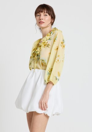 Femme portant un chemisier jaune à volants fleuri avec des manches trois-quarts et une jupe boule blanche, posant devant un fond uni.