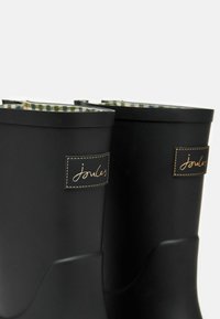 Must värvilised kummisaapad, millel on õmmeldud brändi silt, rohelise šoti mustriga sisepinnal ja sile, ümar ülemine äär.