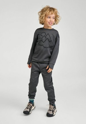 Bambino con capelli biondi ricci che indossa una felpa grigio scuro con la scritta "move mountains" e pantaloni abbinati, in piedi con la testa leggermente girata a destra.