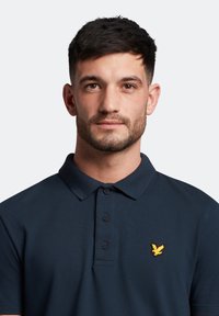 Polo shirt blu navy con colletto, cinque bottoni neri e un piccolo logo ricamato di un uccellino giallo sul petto sinistro.