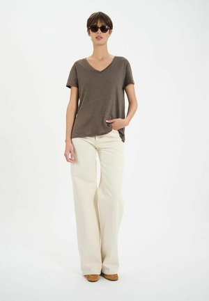 T-shirt vert olive à col en V associé à un pantalon large crème. Le modèle porte des lunettes de soleil sombres et a les cheveux courts, se tenant devant un fond blanc.