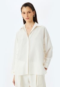 Camisa blanca de manga larga con una sutil textura a rayas, puños abotonados y cuello. Presenta botones dorados en el frontal y en las mangas.