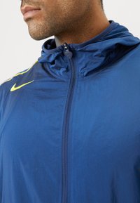 Blå nylonjacka med dragkedja fram och huva. Har en gul Nike-logotyp på vänster sida och en slät, lätt textur.