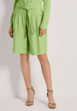 Bermudas en lin de couleur vert clair avec des plis sur le devant, une ceinture à nouer et des poches, associées à des sandales à talons hauts à lanières de couleur vert olive.
