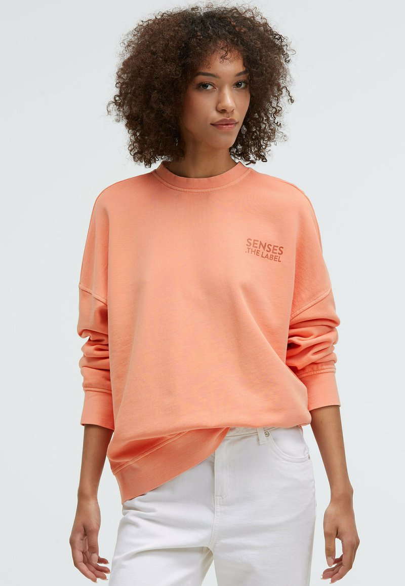 Peach-colored oversized sweatshirt with crew neck, dropped shoulders, and "SENSES THE LABEL" printed in light gray on the chest.