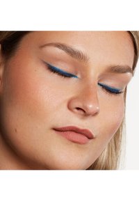 Närbild av ett ansikte med blå eyeliner som framhäver stängda ögon, slät hud med en naturlig ton och matta, nude läppar.